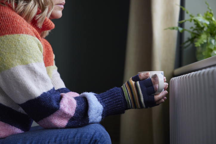 Eine Frau sitzt im Pullover und mit Handschuhen mit einer Tasse in der Hand vor einem Heizkörper.